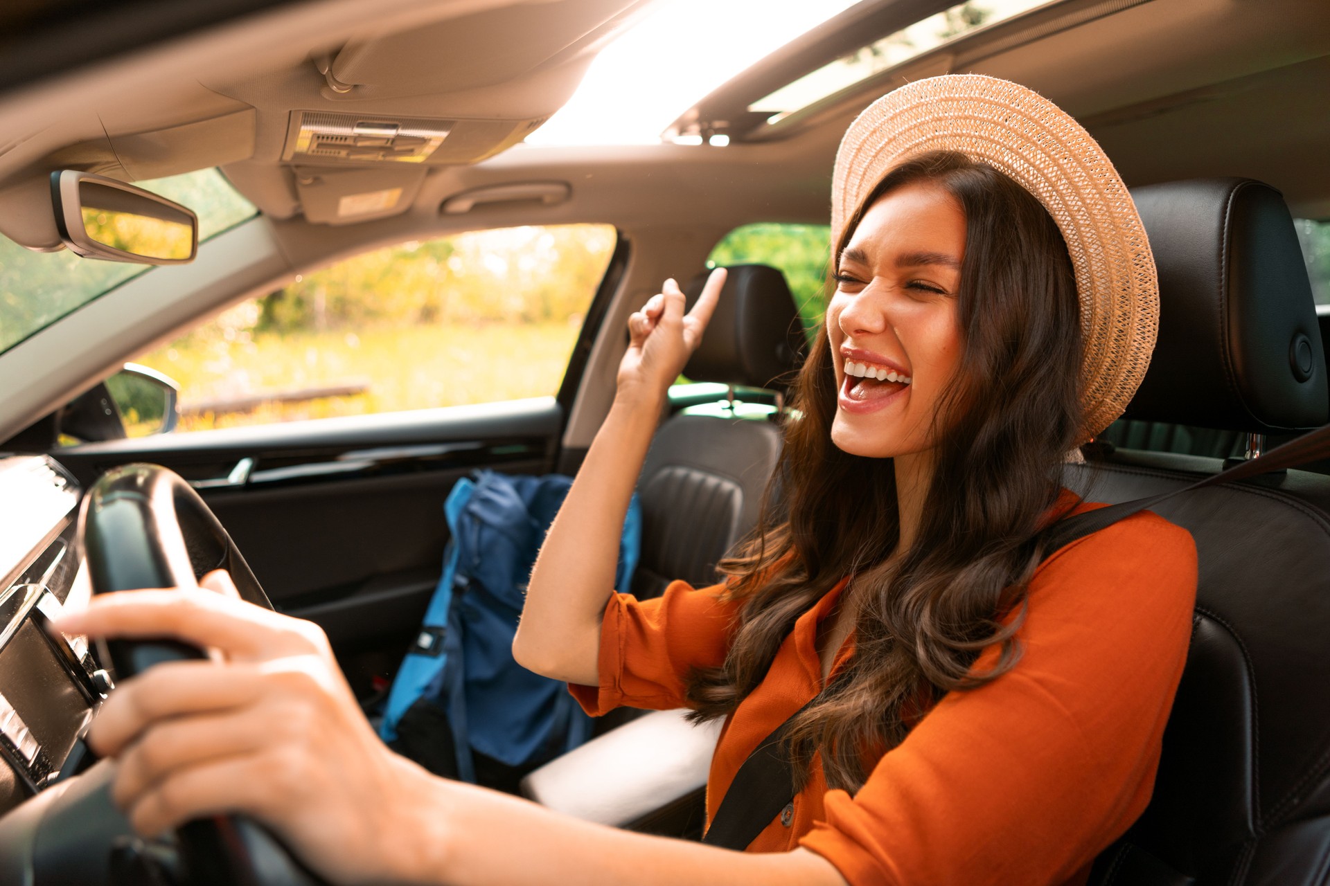 Overjoyed woman tourist enjoying car ride alone, listening to music, singing songs and dancing while driving auto with backpack nearby on passenger seat